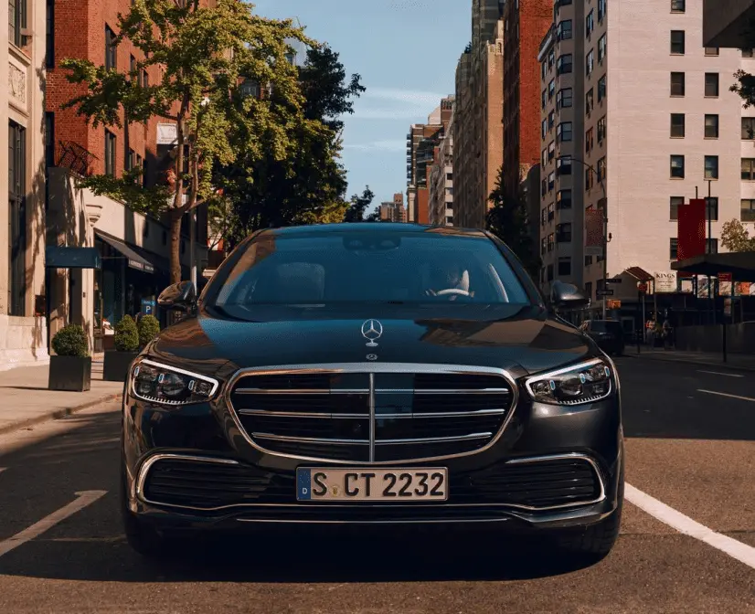 Vue de face d'une voiture noire Mercedes-Benz garée sur une rue urbaine bordée d'immeubles et d'arbres.
