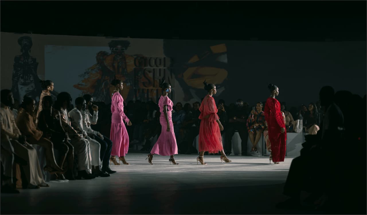 Défilé de mode avec quatre mannequins portant des robes élégantes en rouge et rose, marchant sur un podium face à un public assis.