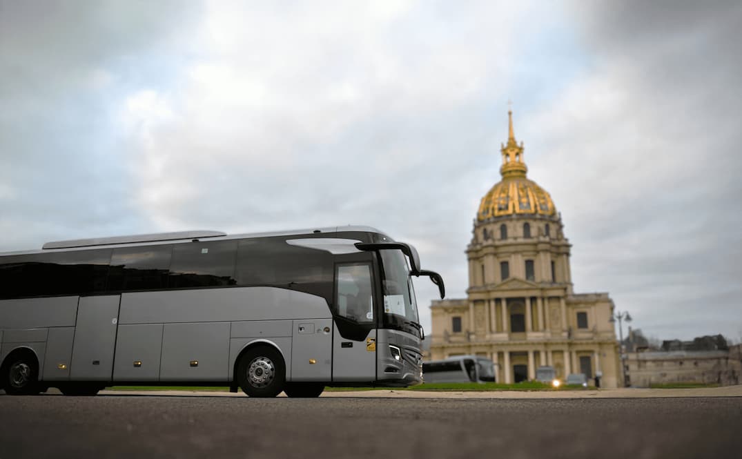 Autocar gris garé devant un bâtiment historique avec un dôme doré sous un ciel nuageux.