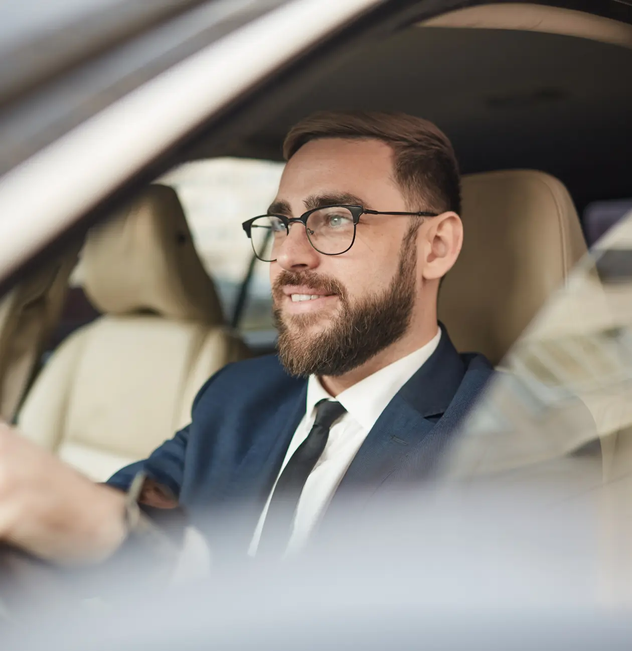 Homme souriant portant des lunettes, costume bleu et cravate noire, assis dans une voiture au siège beige.