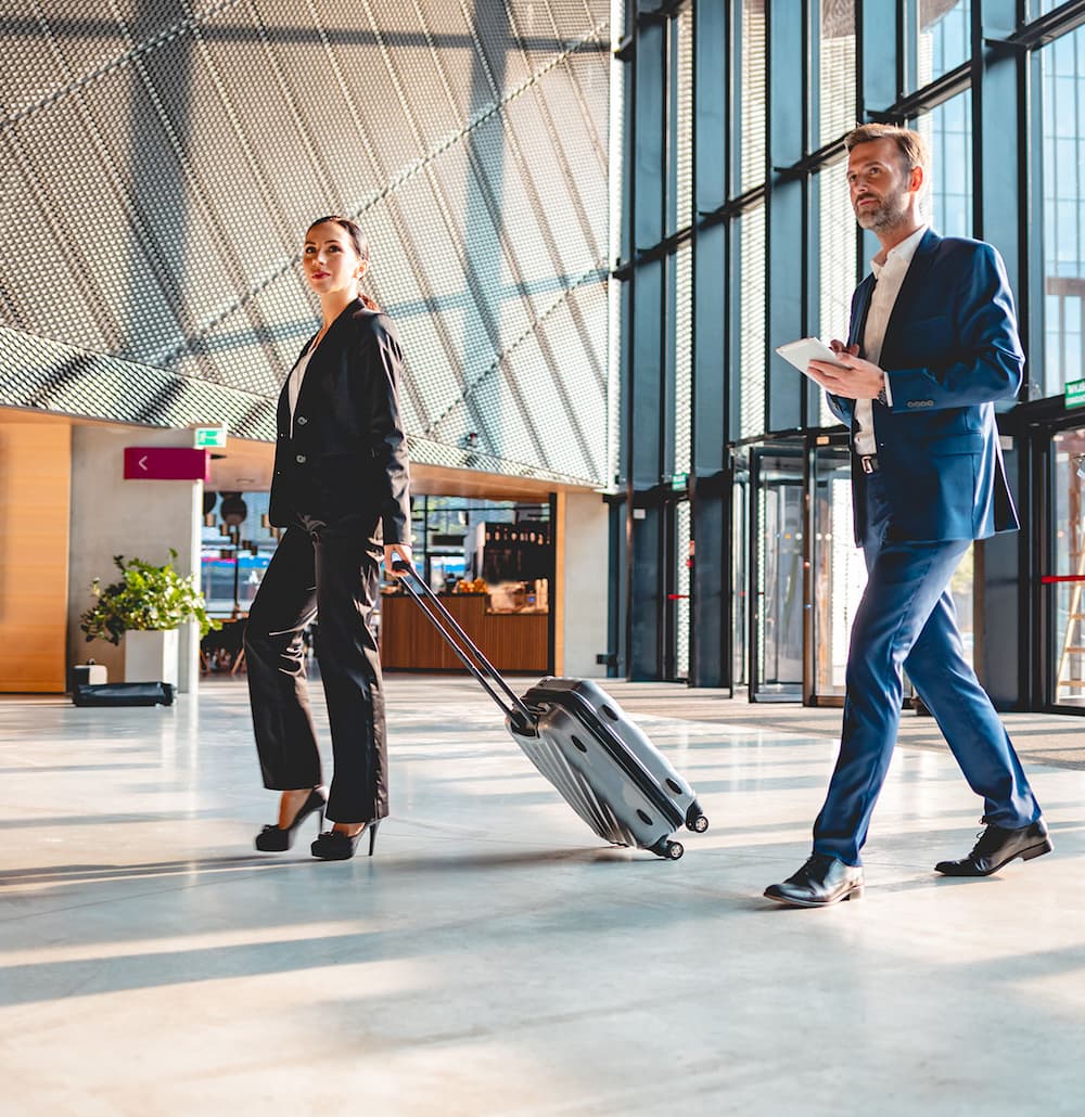 Une femme en tailleur noir tire une valise à roulettes dans un hall d'aéroport moderne, suivie d'un homme en costume bleu tenant une tablette.