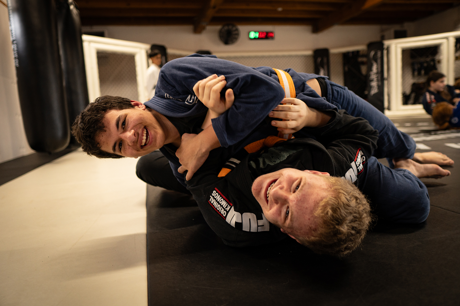 Zwei Jugendliche lachen beim Kampfsporttraining auf der Mattenfläche.