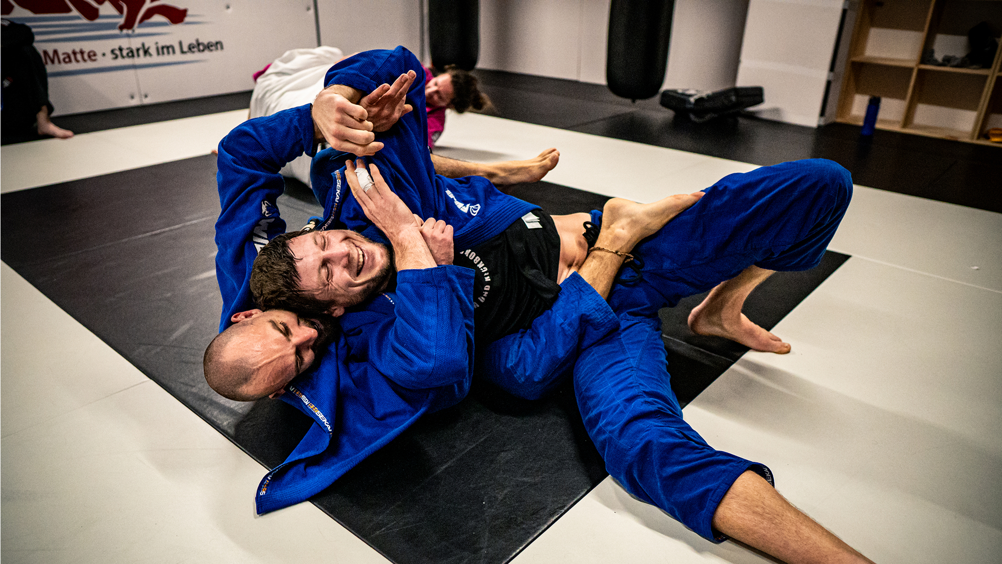 Zwei Männer lachen und trainieren Brazilian Jiu Jitsu Techniken am Boden.