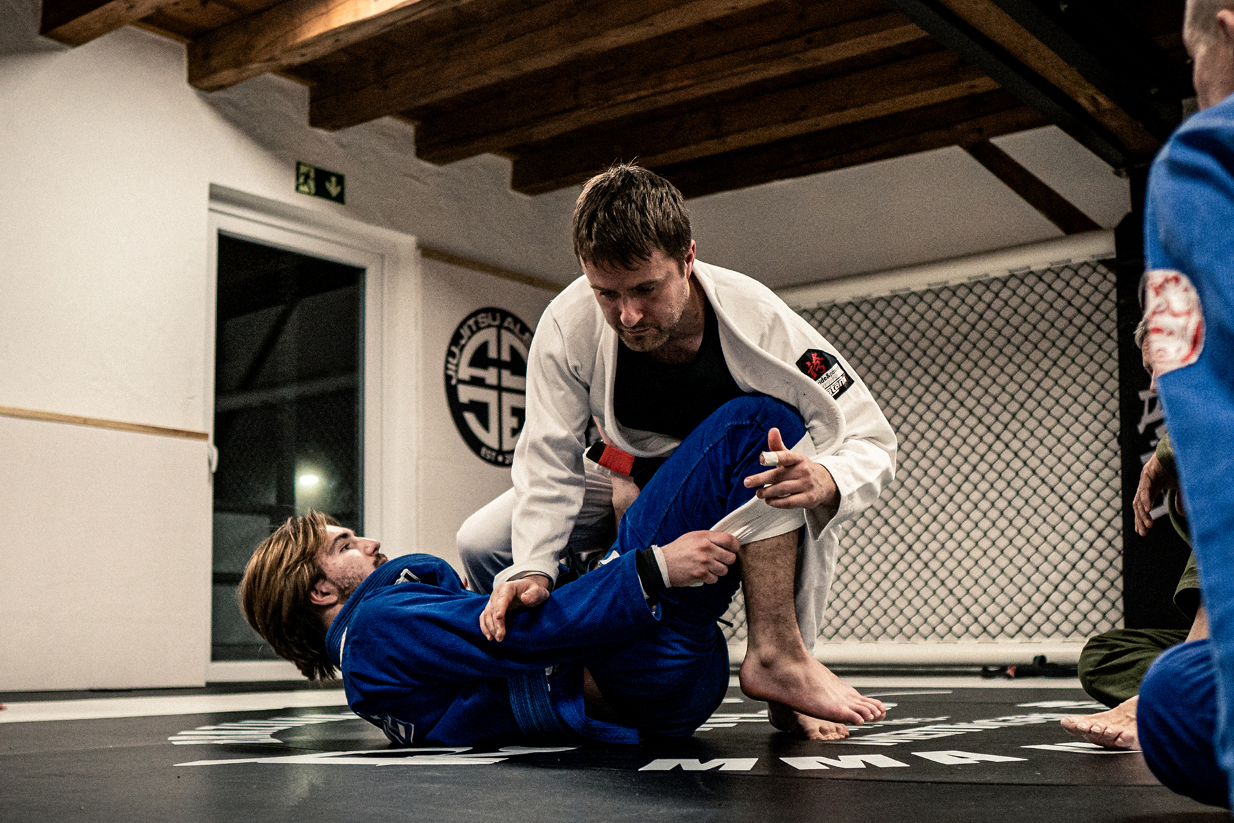 Zwei Männer in Jiu-Jitsu-Anzügen trainieren am Boden, umgeben von anderen Beobachtern auf der Matte.