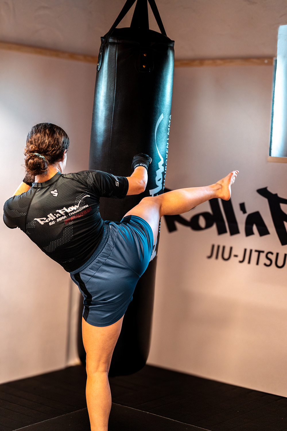 Person trainiert Muay Thai mit einem hohen Kick gegen einen schwarzen Sandsack im Gym.