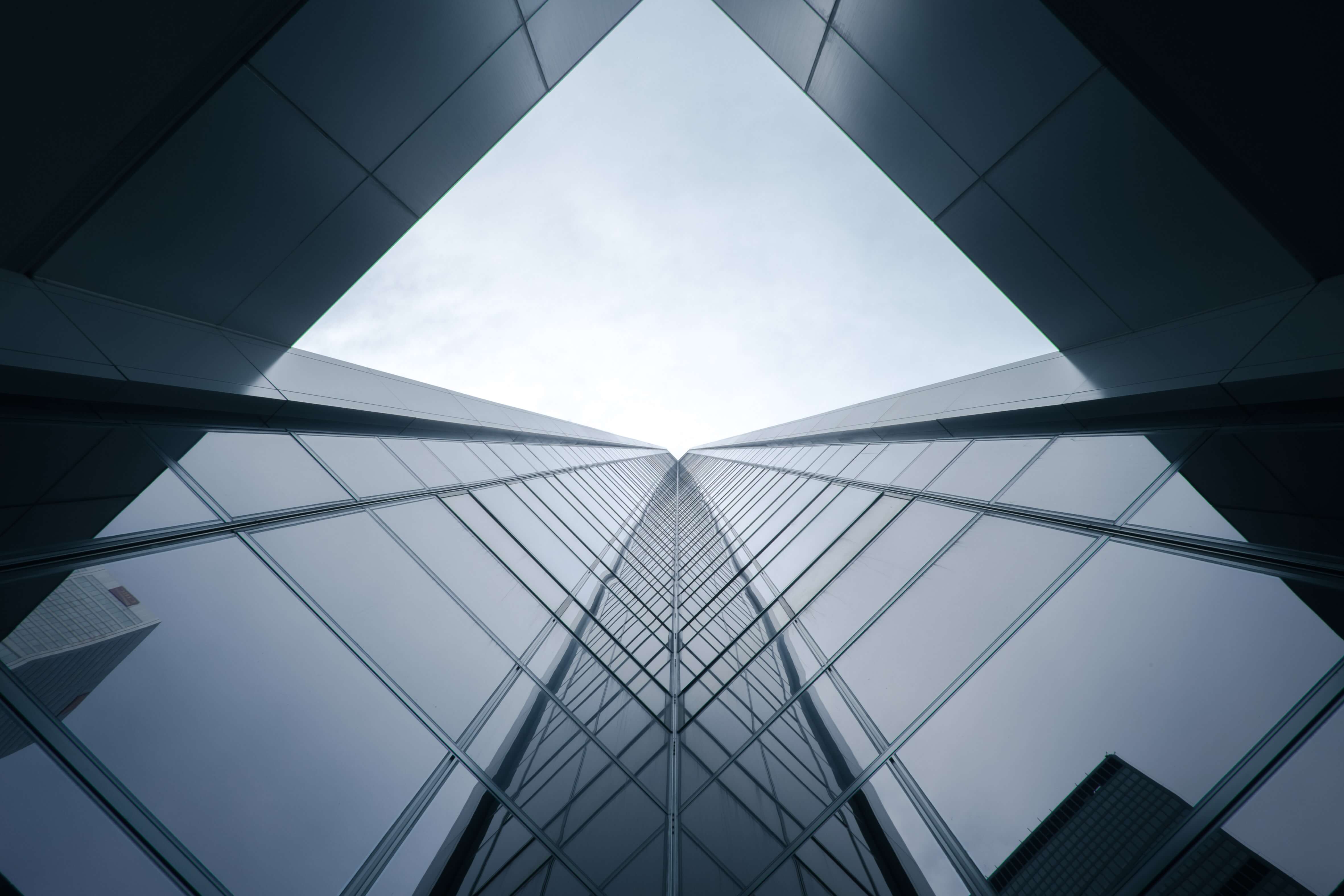 Looking up at a modern glass skyscraper with reflections of nearby buildings and a cloudy sky.