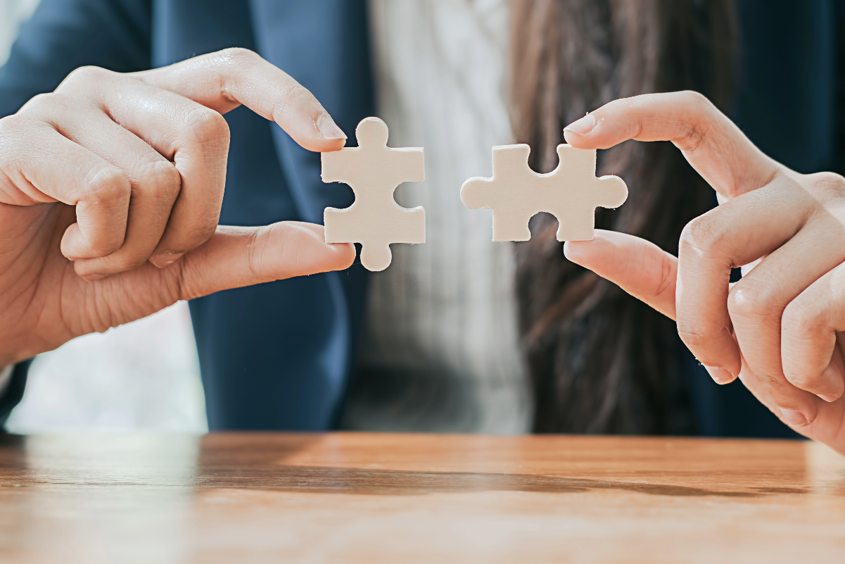 Two hands holding two beige puzzle pieces about to be connected over a wooden table.