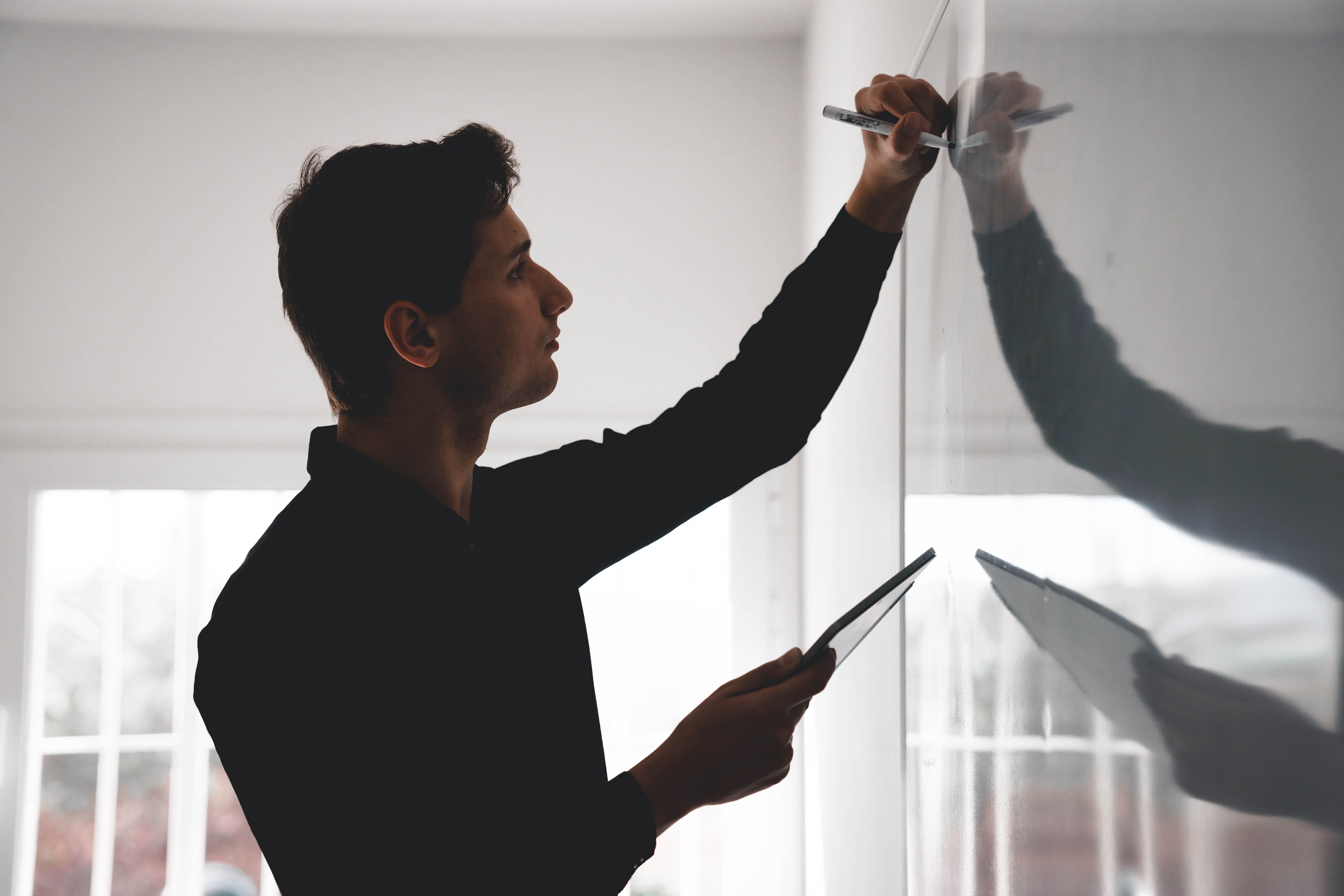 Man holding a tablet writing on a whiteboard with a marker in a bright room.