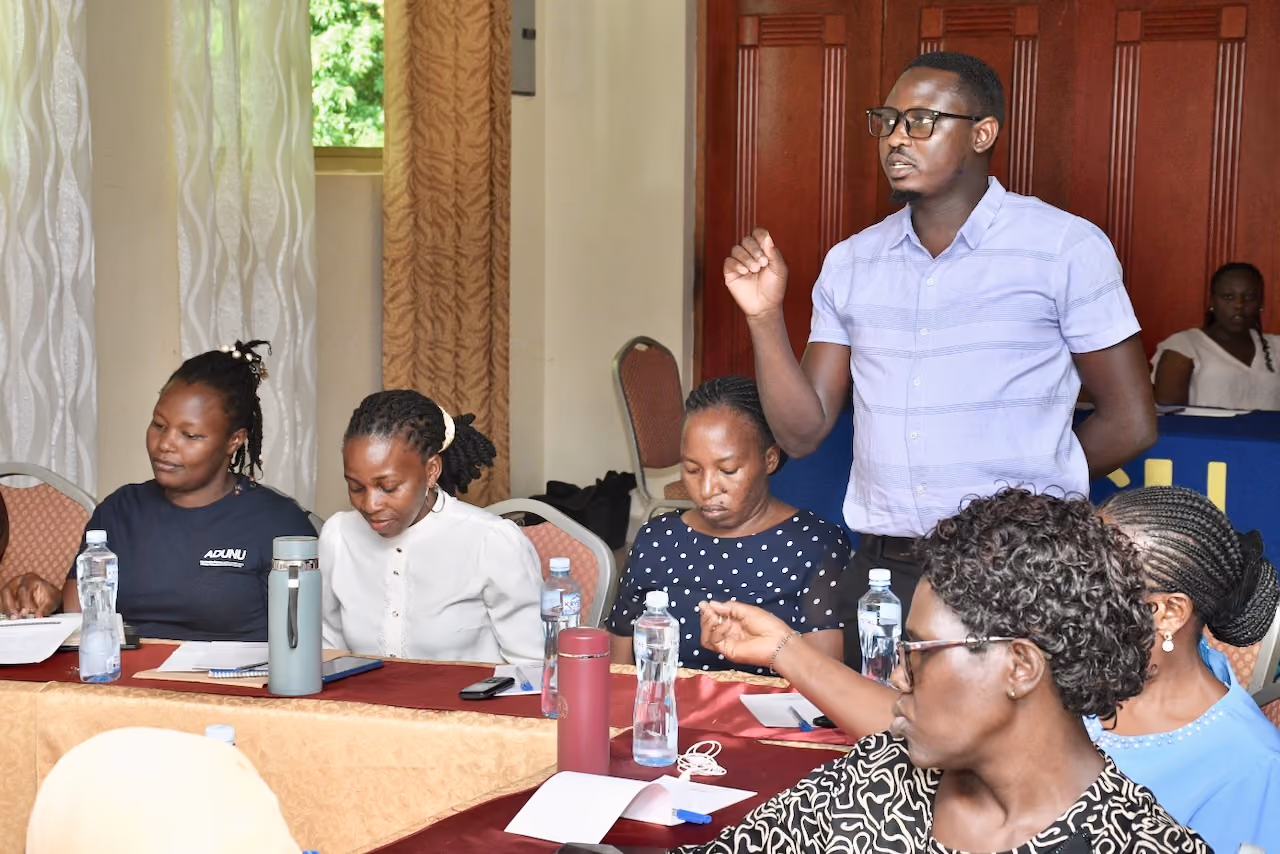 Group of adults in a meeting with one man standing and speaking while others listen and take notes.