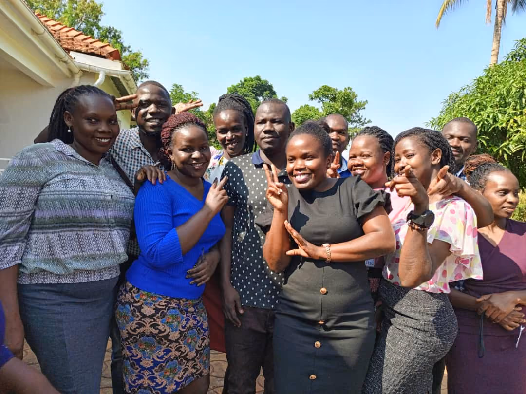 Group of smiling men and women posing outdoors with some making peace signs and pointing.