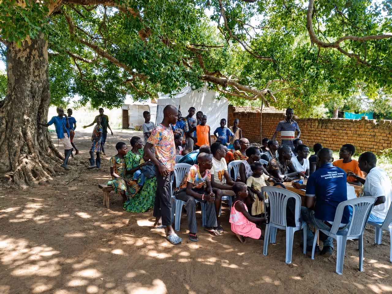 Community gathering with men, women, and children sitting and standing under a large leafy tree.