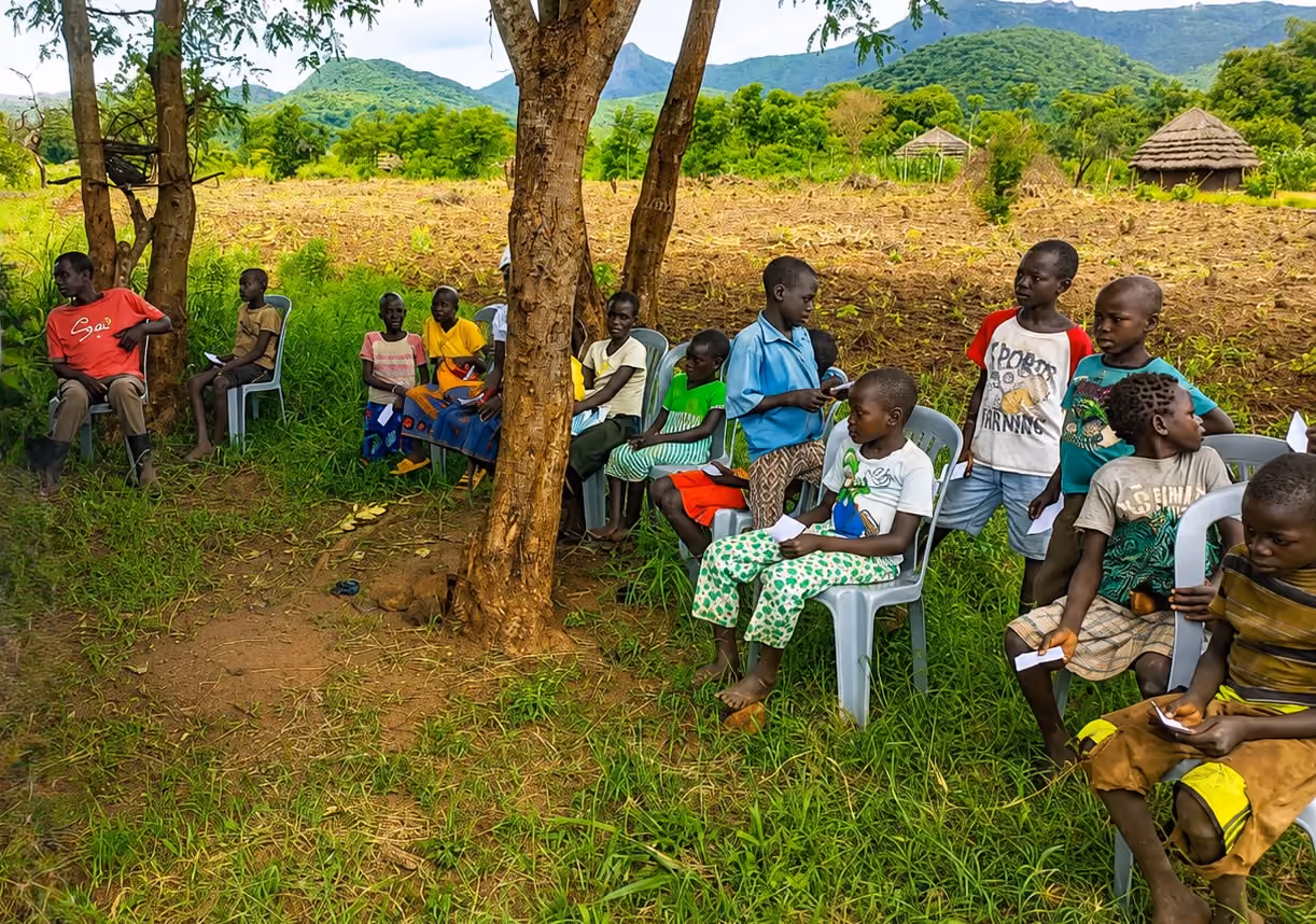 Children sitting outside under trees on plastic chairs with rural farmland and huts in the background.