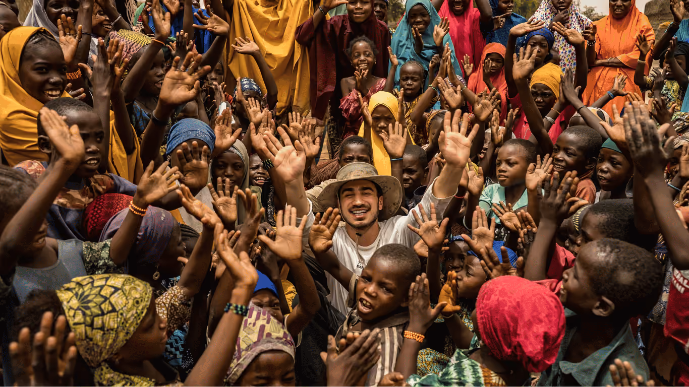 Smiling man wearing a hat surrounded by joyful children raising their hands in colorful clothes outdoors.