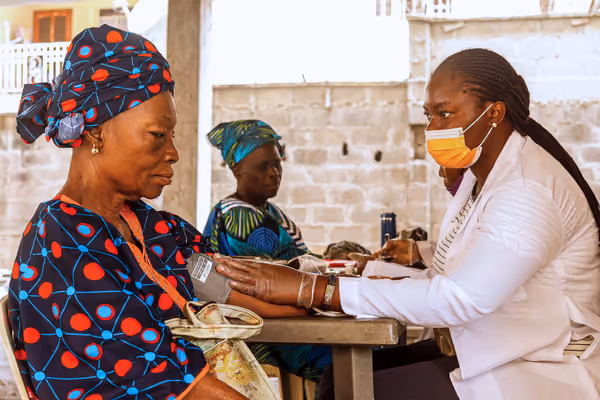 Healthcare worker wearing mask checks blood pressure of an elderly woman in colorful traditional clothing.