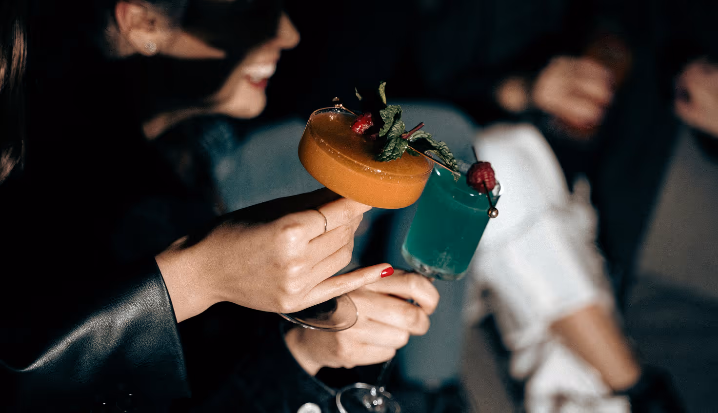 Image from Unsplash - Two people clinking colorful cocktails garnished with berries and mint in a dim setting.