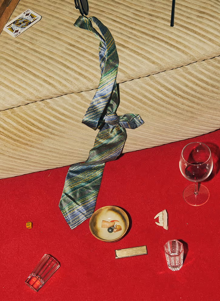 Image from Death to Stock - Patterned necktie on a beige couch and red carpet with a wine glass, dice, cards, and small objects.