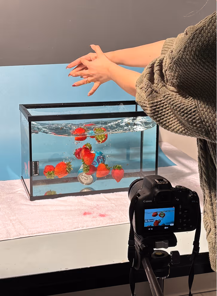 Photographed by Xhalia Martineau - Person prepares to photograph strawberries and soda cans splashing in a glass tank of water.