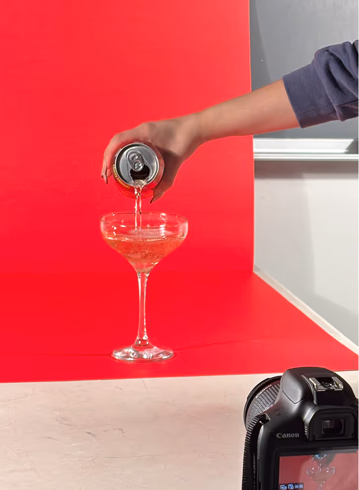 Photographed by Xhalia Martineau - Hand pouring a drink from a can into a coupe glass against a red backdrop with a camera in front.