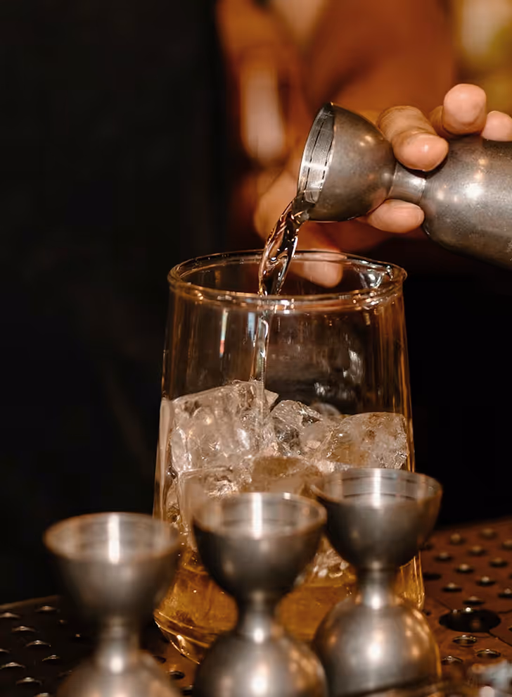 Image from Unsplash - Hand pouring liquid from a metal jigger into a glass filled with ice cubes on a bar counter.