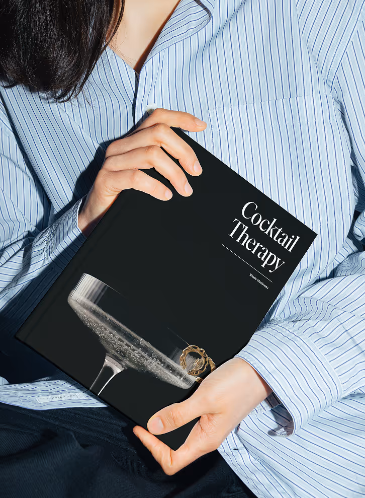 Mockup from Maison Mockup - Person in striped shirt holding a black book titled 'Cocktail Therapy' with a champagne glass image.