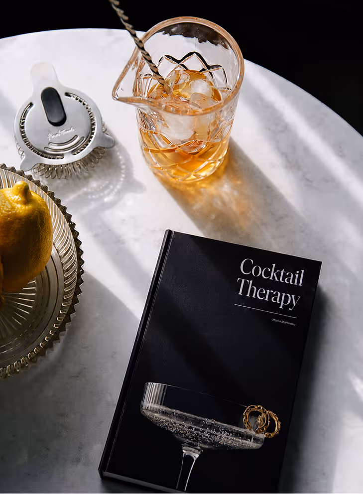AI Generated image by ChatGpt - Cocktail Therapy book on a table next to a glass with a drink and ice, a lemon, and a strainer.