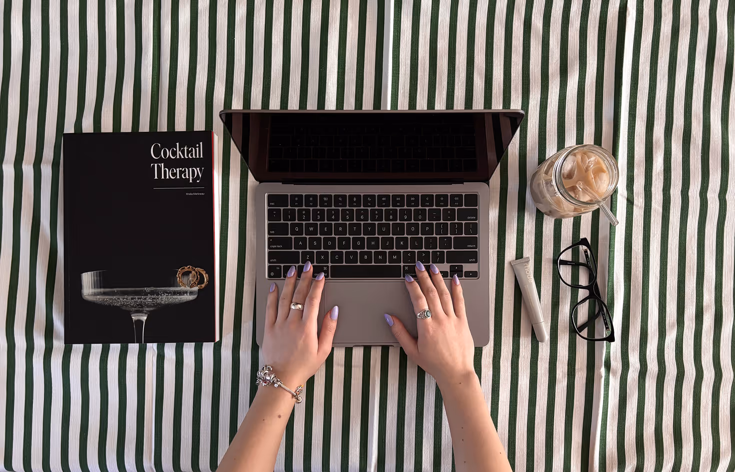 Photographed by Xhalia Martineau - Hands with lavender nails typing on laptop keyboard on green and white striped cloth with book and iced coffee.