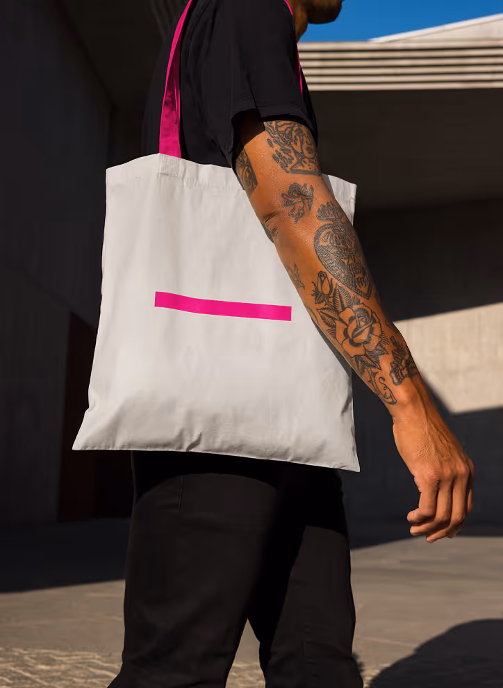 Mockup from Maison Mockup - Person with tattooed arm wearing black clothes carrying a white tote bag with pink handles and design.