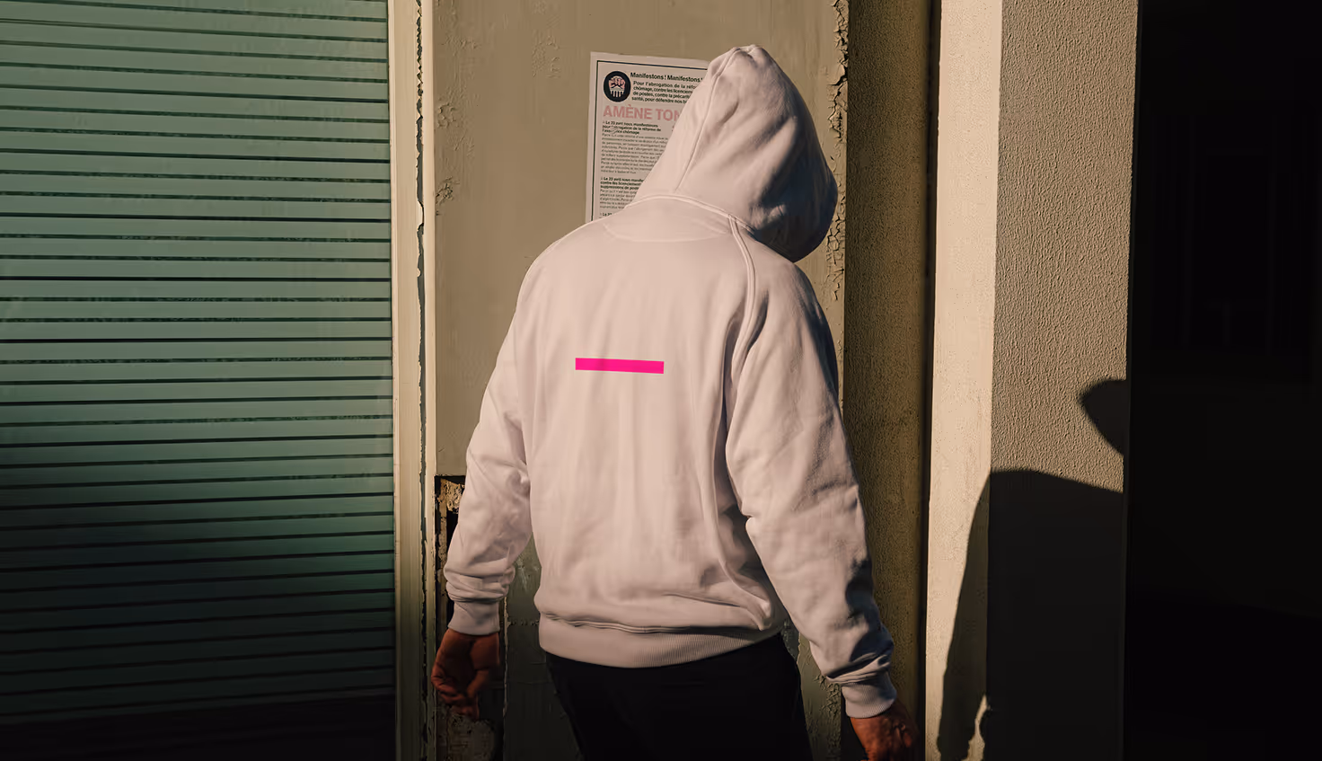 Mockup from Maison Mockup - Person wearing a white hoodie with the hood up standing in front of a wall with a poster.