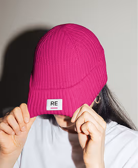 Mockup from Maison Mockup - Person adjusting bright pink knit beanie with 'RE' patch, face partially covered.
