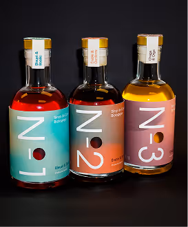 Photographed by Xhalia Martineau - Three glass bottles with labels L-1, N-2, N-3 containing colored liquids on a black background.