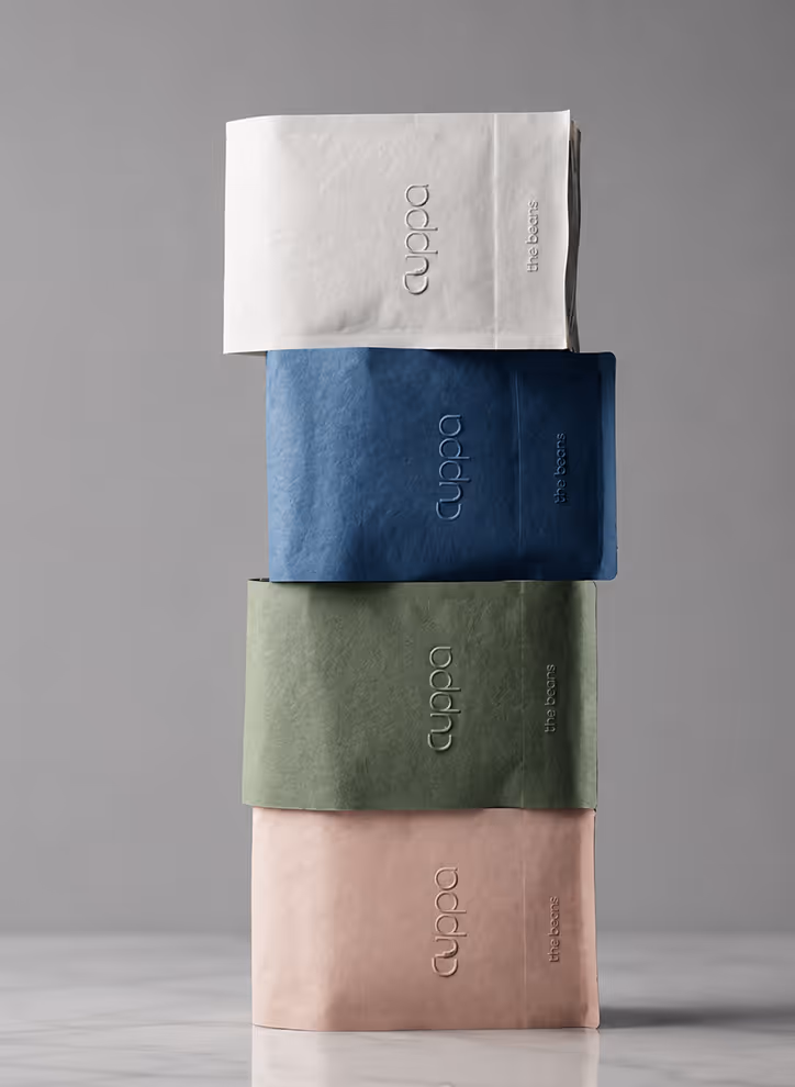 AI Generated image with ChatGpt -  Four stacked coffee bags in white, blue, green, and pink with embossed 'cuppa' and 'the beans' text.