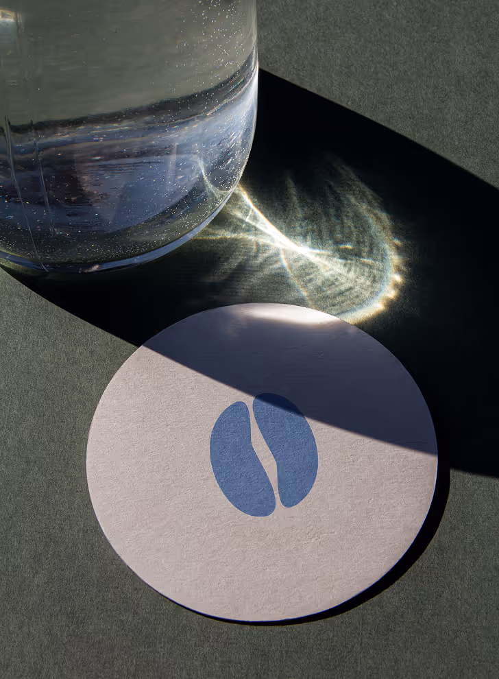 Mockup from Maison Mockup - Glass container casting light reflections above a round coaster with two blue coffee bean shapes.