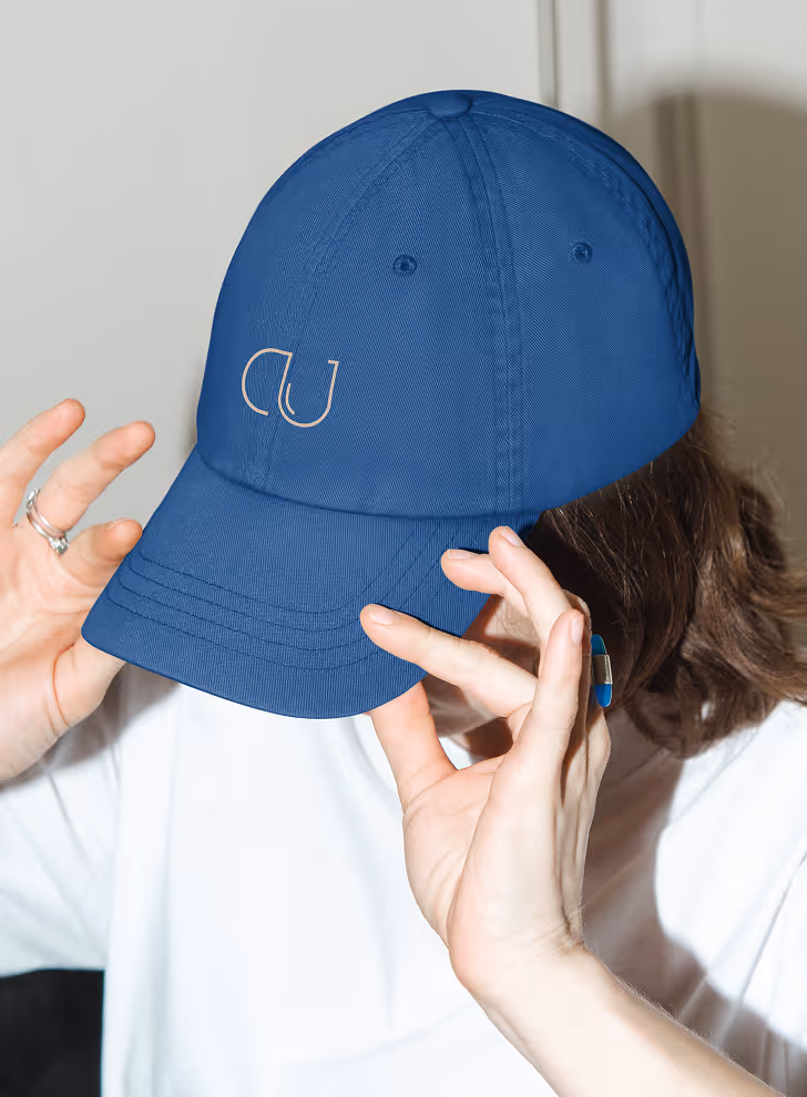 Mockup from Maison Mockup - Person adjusting a blue baseball cap with a white stylized letter design on the front.