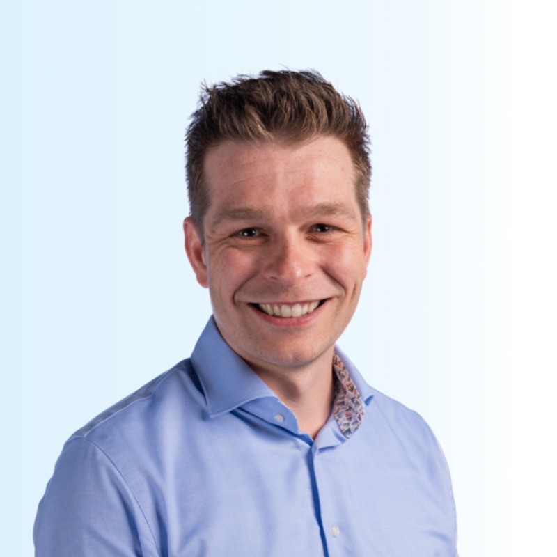 Portrait of a smiling man with short brown hair wearing a light blue dress shirt with a patterned inner collar on a light blue gradient background.