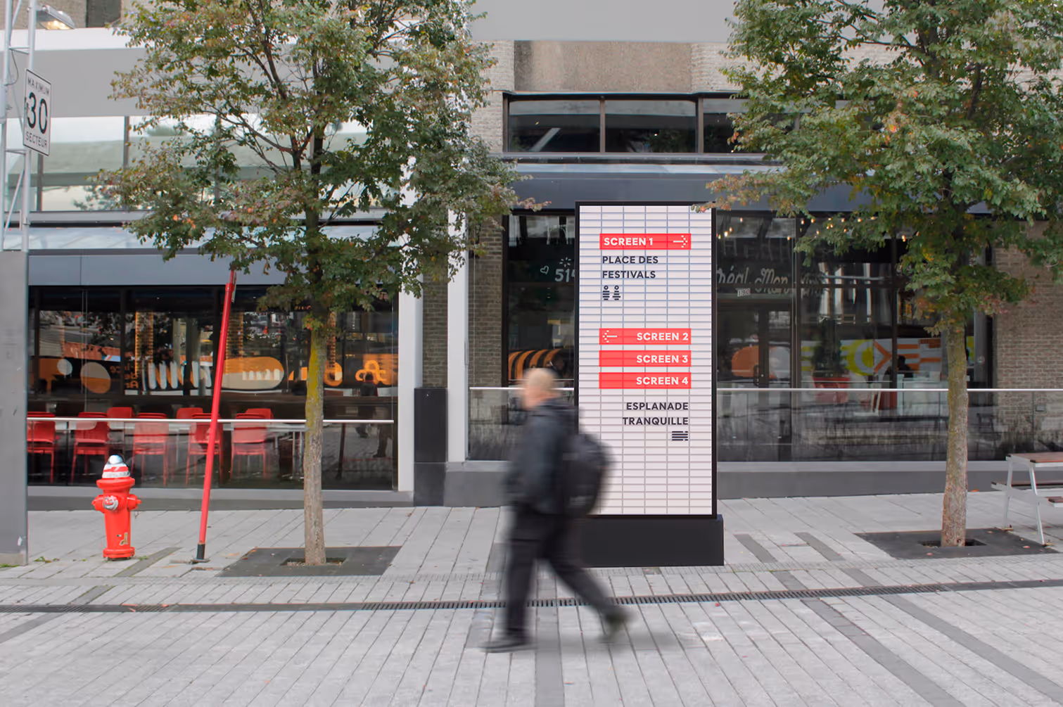 Blurred person walking past a street directory with trees, fire hydrant, and building in background.