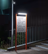 Lit signpost at night with a list of locations near a metal railing and steps.