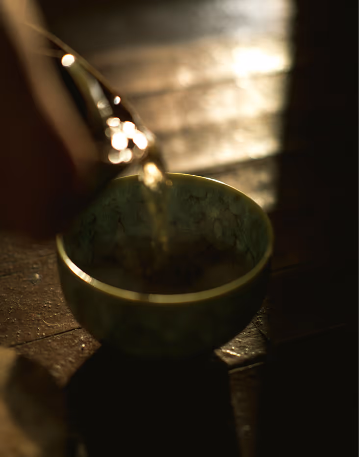 Tea being poured from a glass teapot into a patterned ceramic bowl on a wooden surface.
