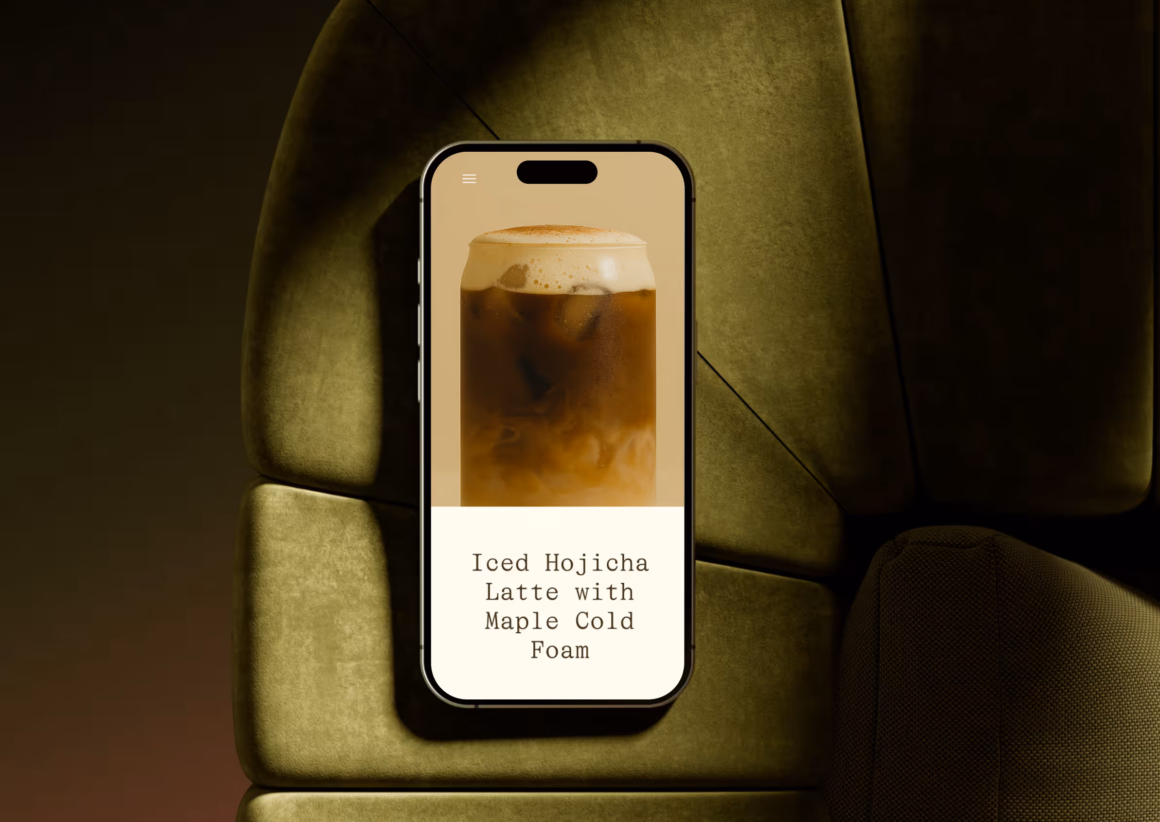 Smartphone displaying an iced Hojicha latte with maple cold foam on a green cushioned surface.