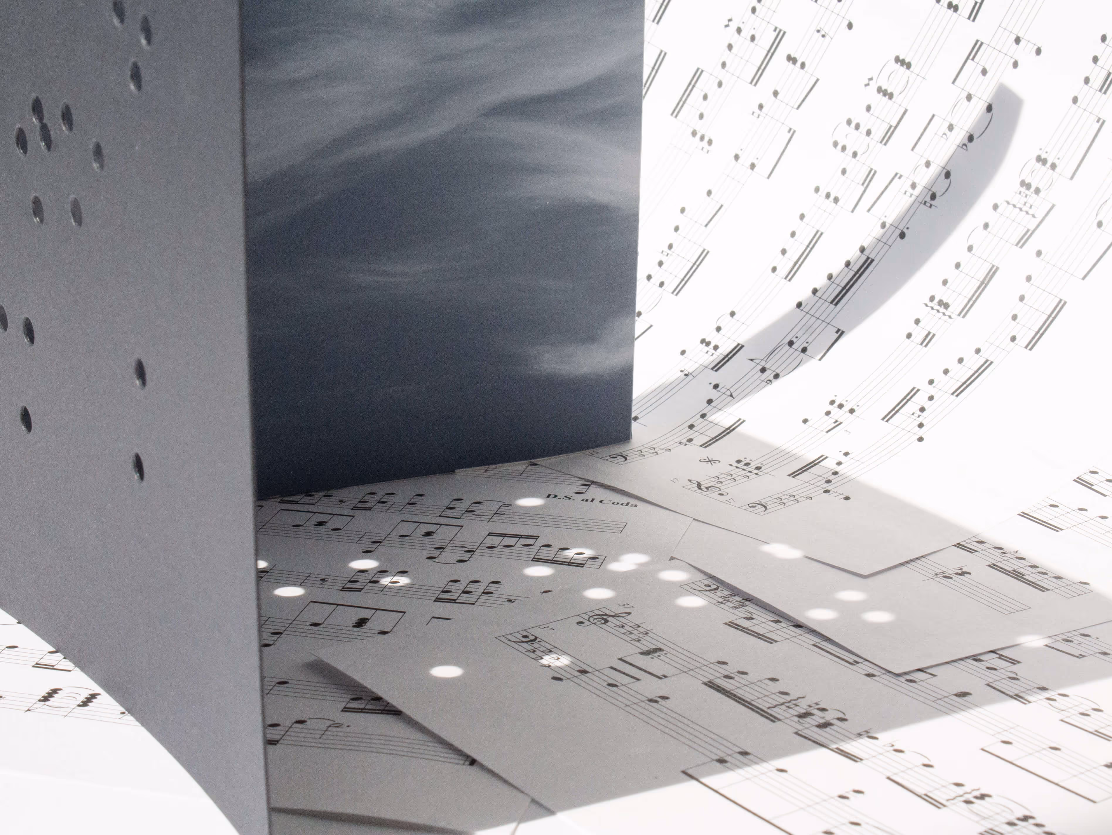 Sheet music pages scattered and bent, with light casting polka dot shadows through a perforated object.