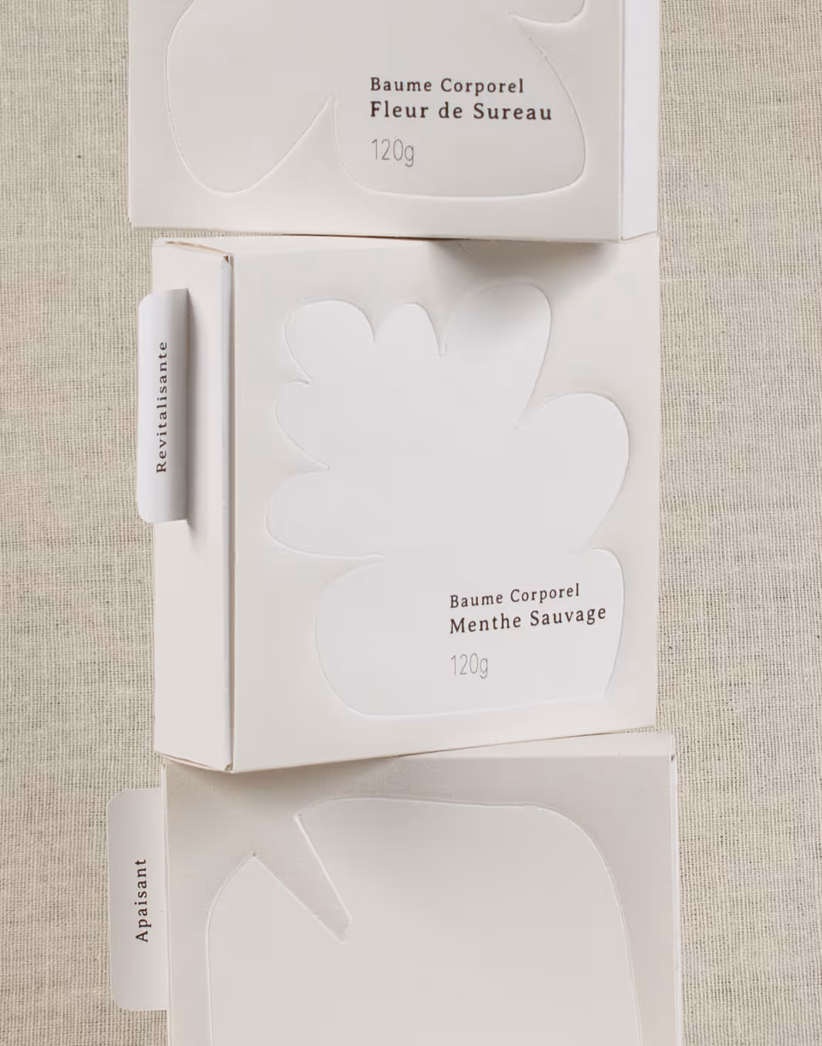 Three white boxes of Baume Corporel body balm in Fleur de Sureau and Menthe Sauvage scents on fabric.