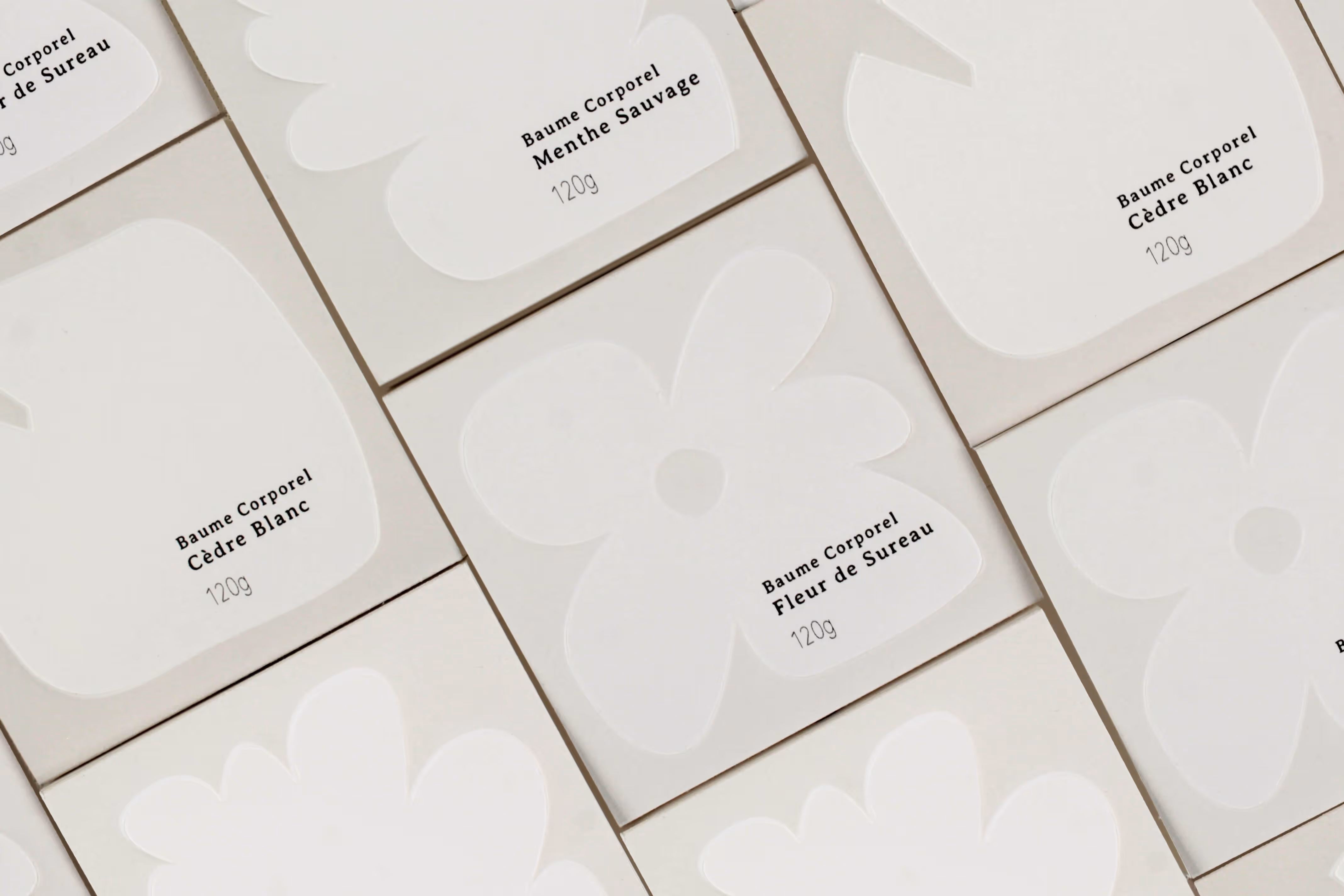 Minimalist white packages of Baume Corporel body balms in various floral shapes and scents arranged flat.
