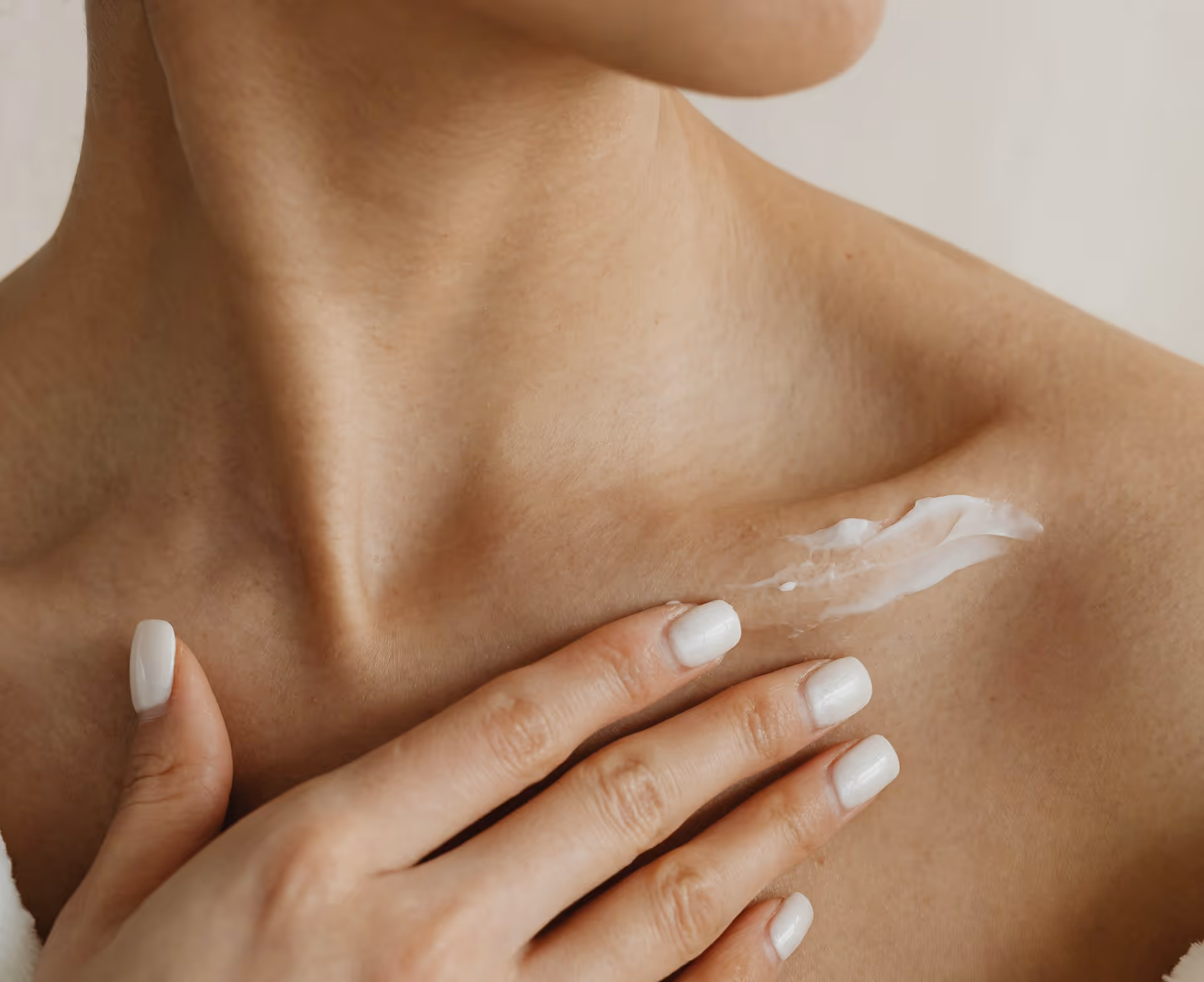 Person with manicured white nails applying white cream to shoulder near collarbone.