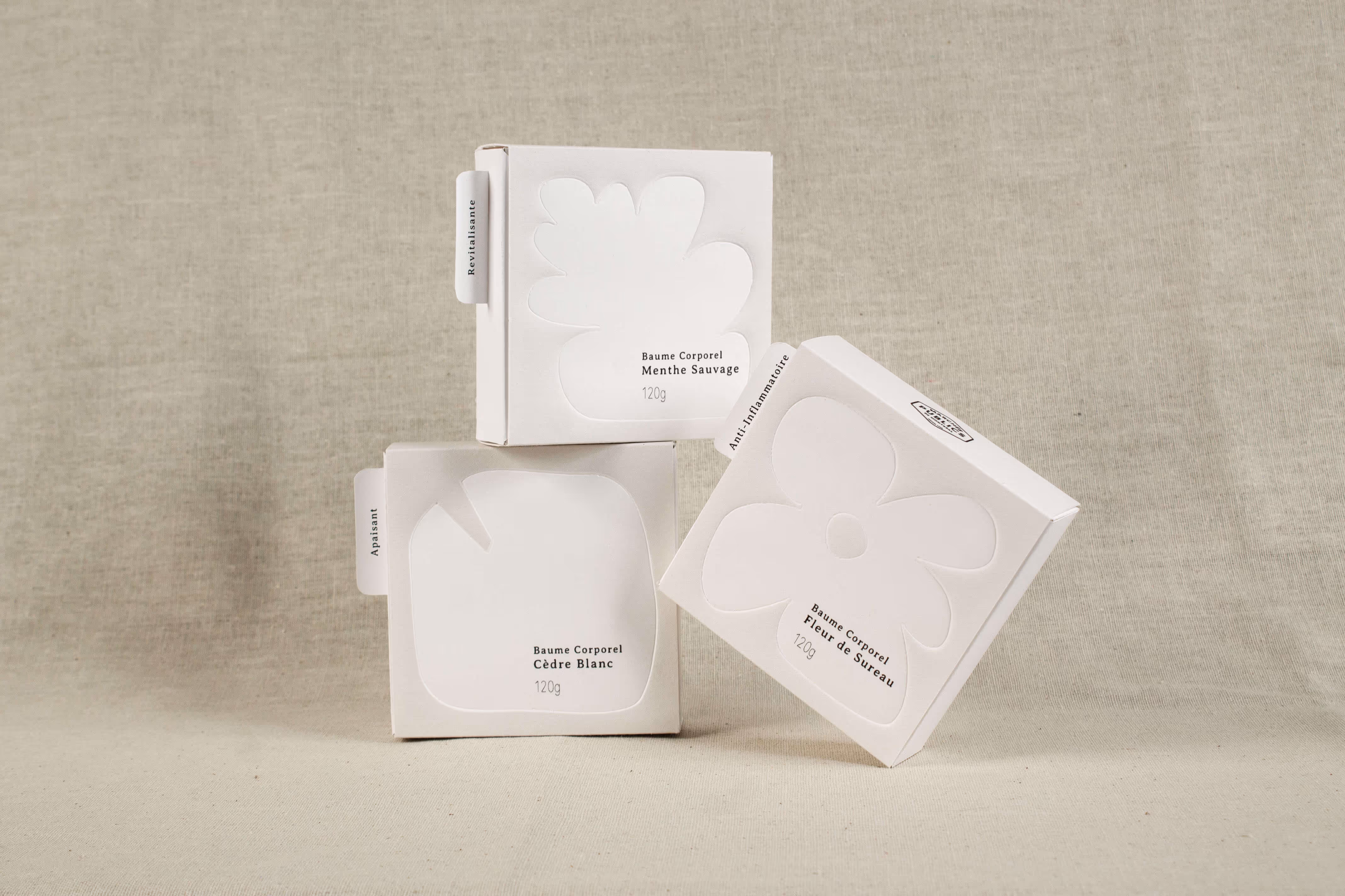 Three white minimalist boxes of Baume Corporel body balm stacked against a beige fabric background.
