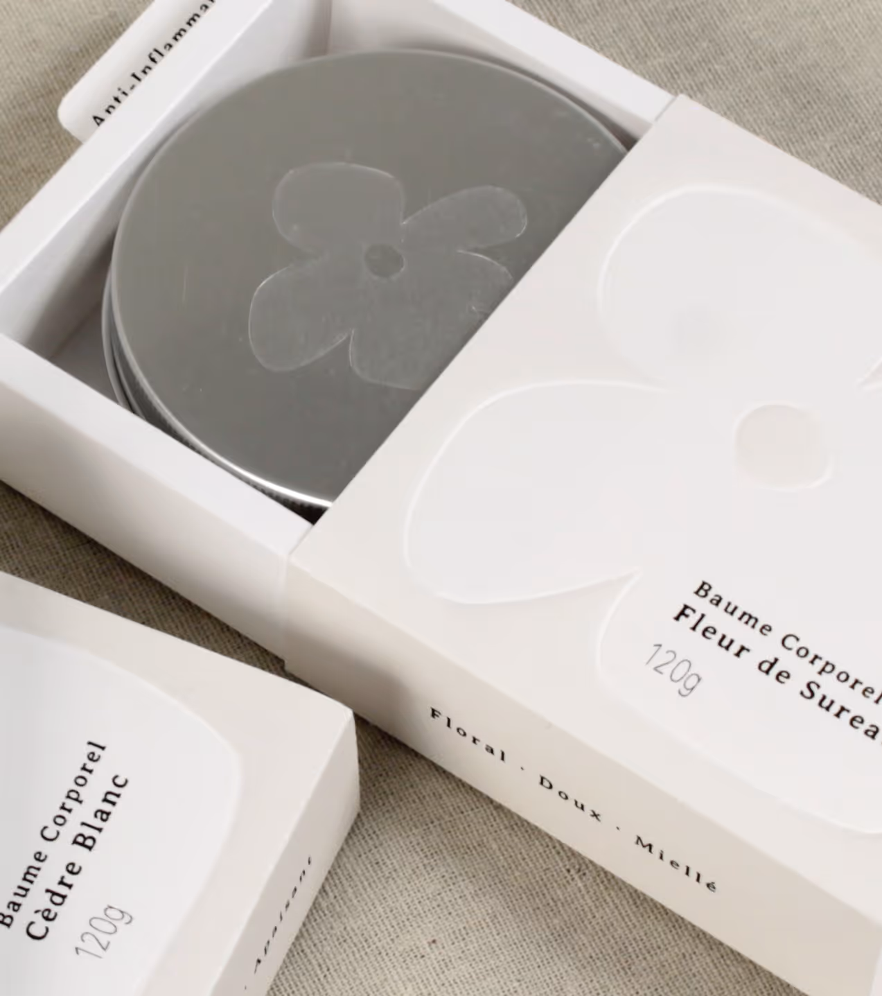 Two white boxes of body balm, one open showing a silver tin with flower design inside.