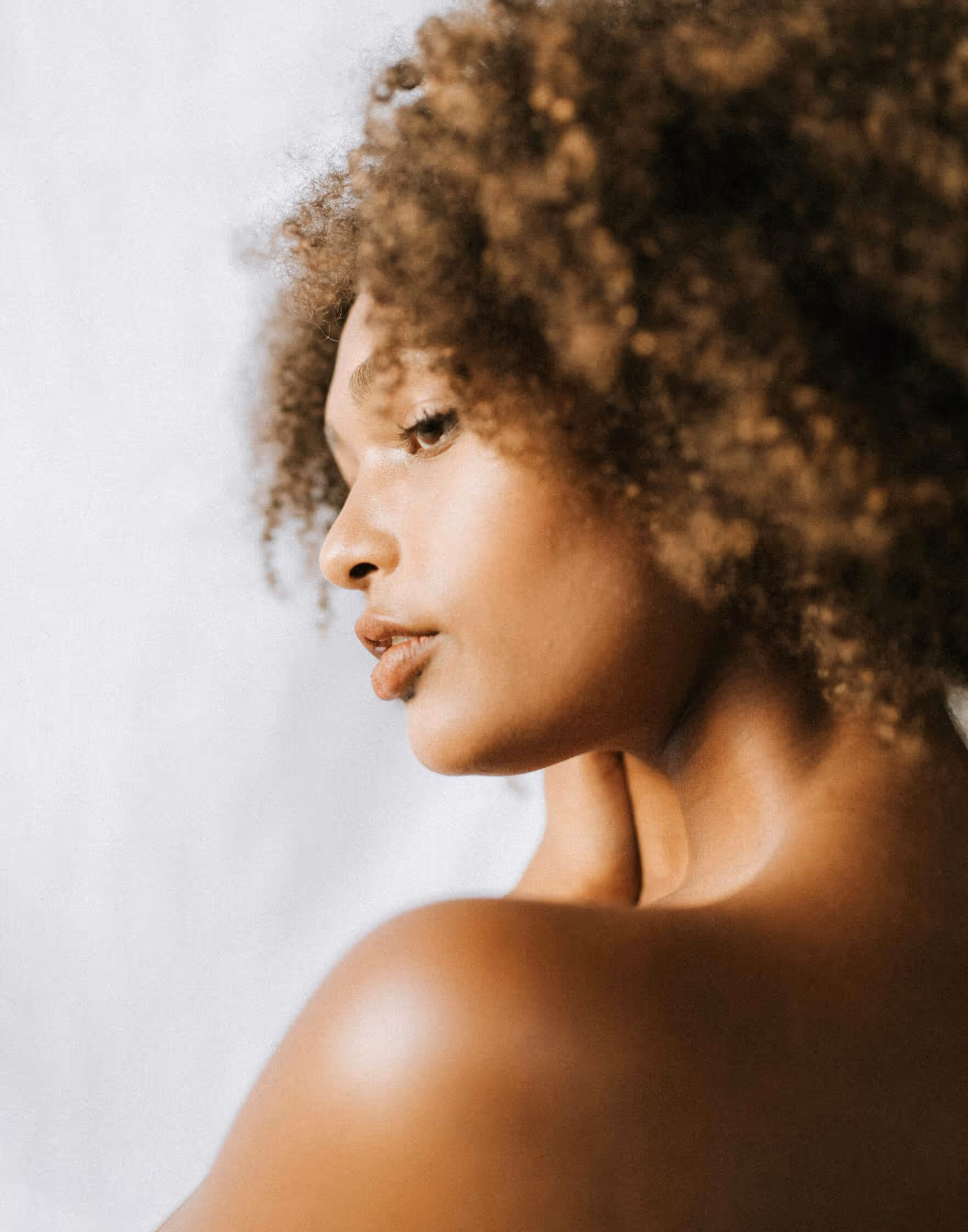 Close-up profile of a woman with curly hair touching her neck against a plain background.