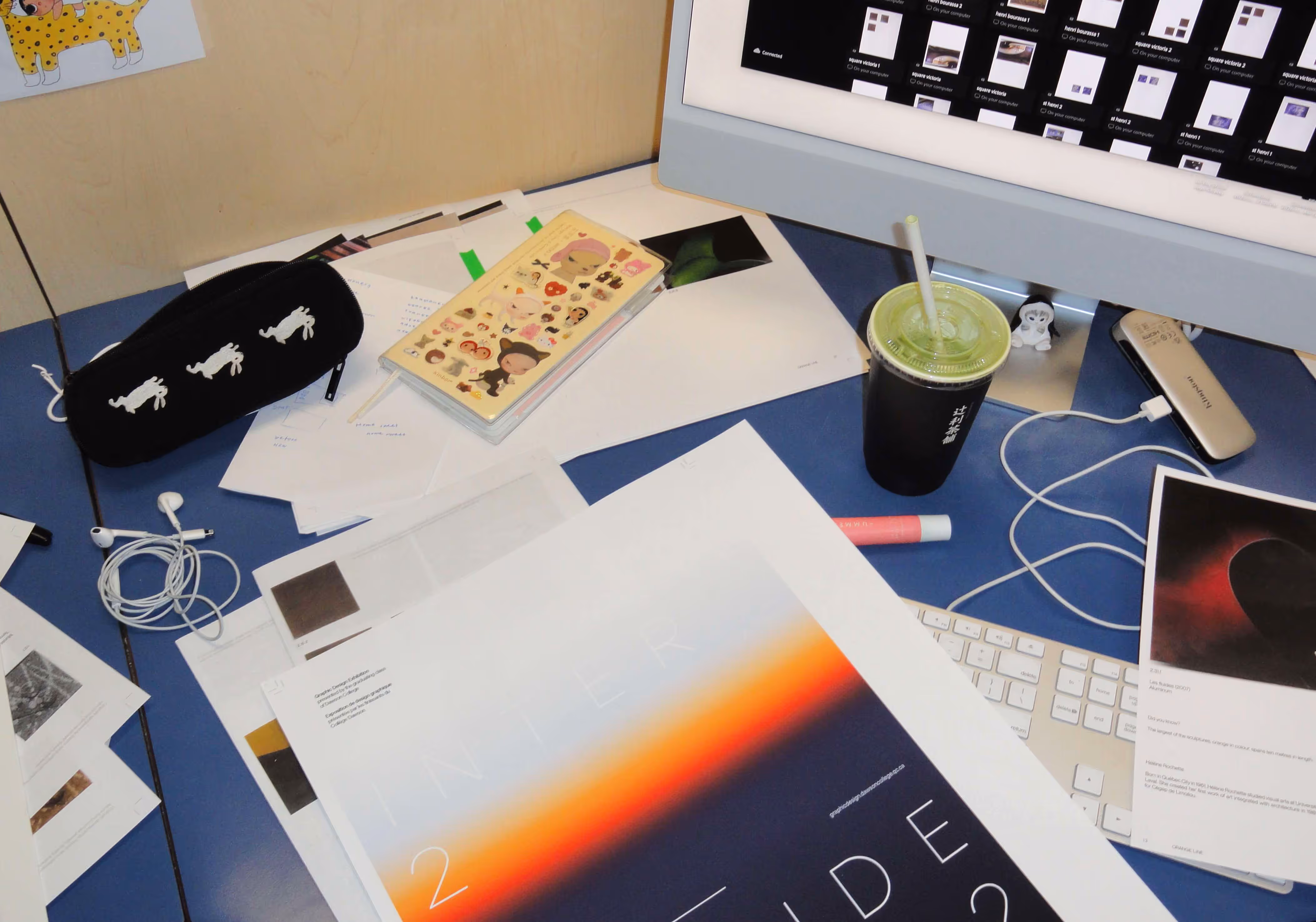 Cluttered desk with papers, stickers, computer, earphones, and a black cup with green drink and straw.