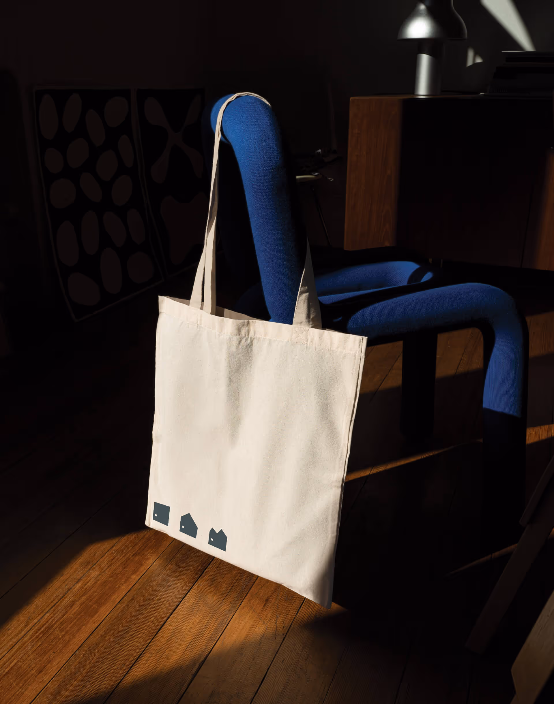 Beige tote bag with small black house icons hanging on blue chair in sunlight on wooden floor.