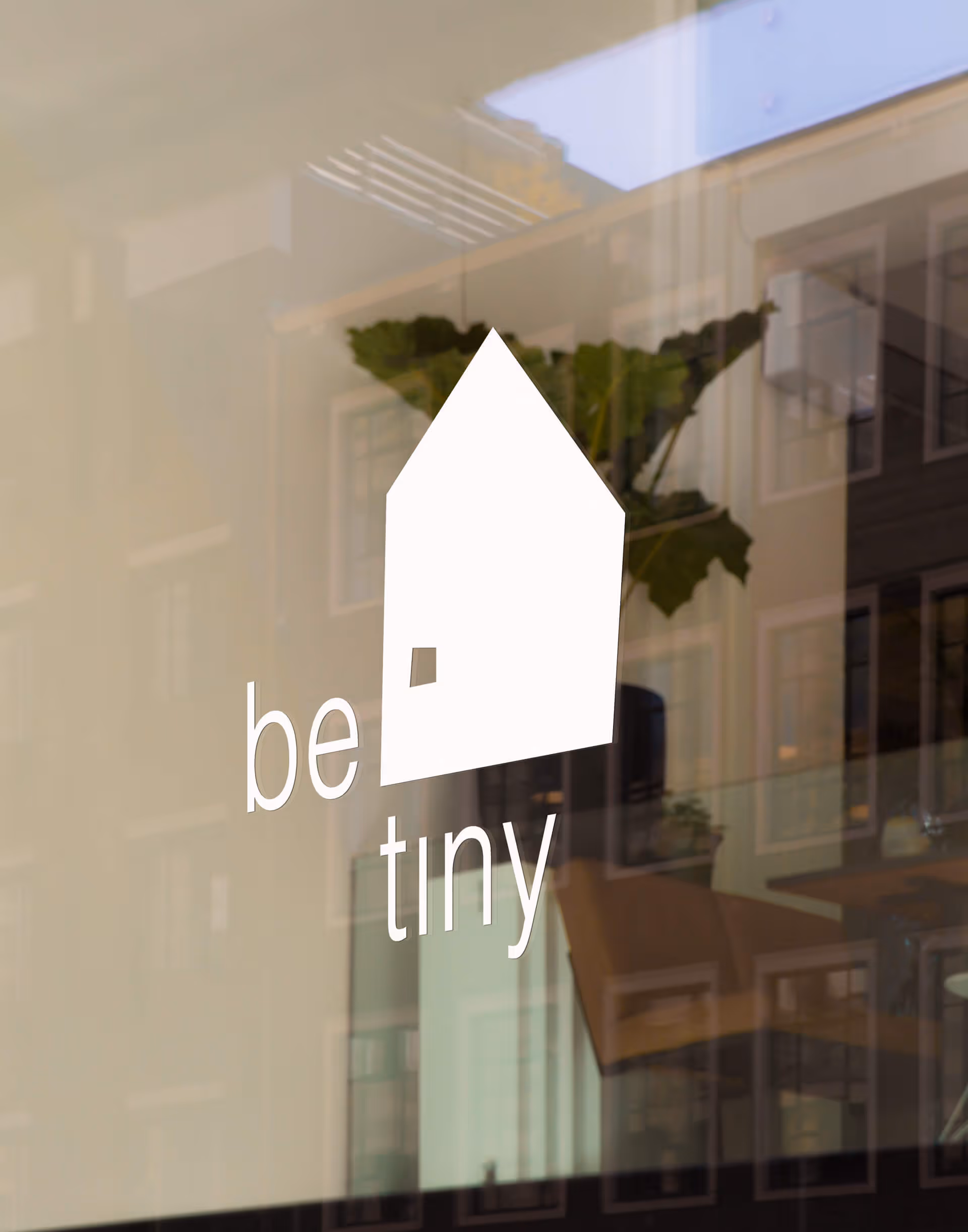 White logo on glass of a simple house shape with the words 'be tiny' inside.