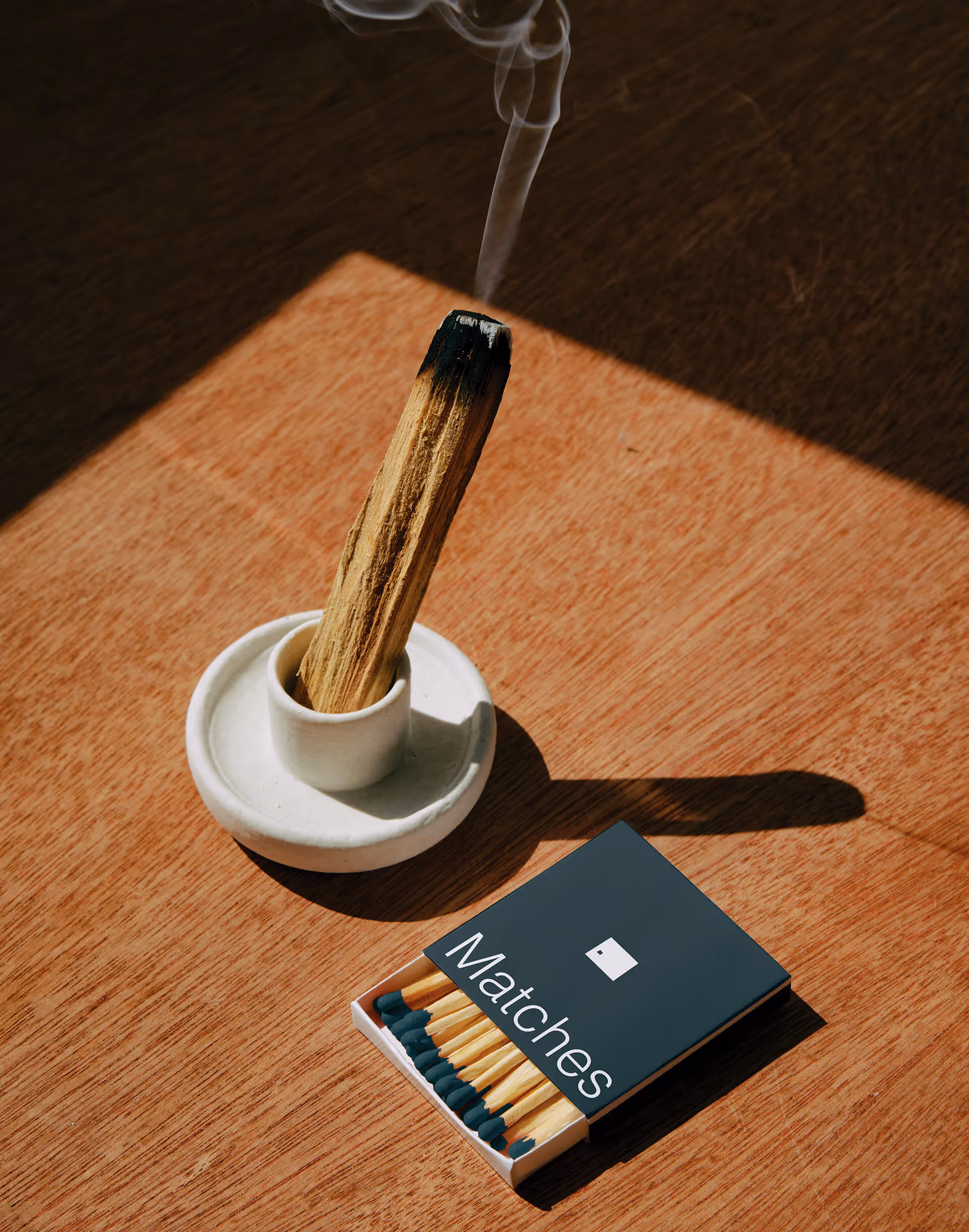 Burning palo santo stick in a white holder beside an open matchbox on a wooden surface.