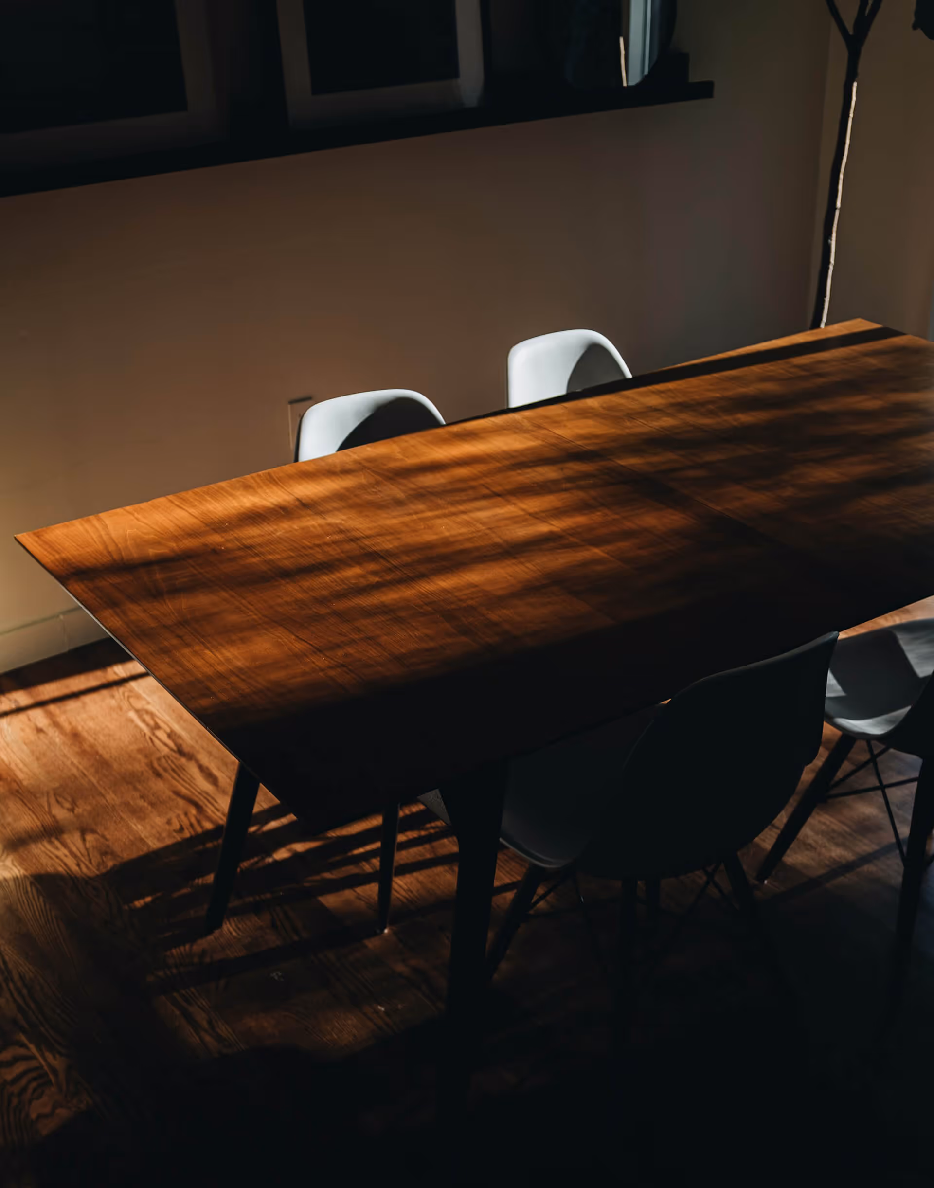 Wooden dining table with white chairs illuminated by sunlight in a dim room.
